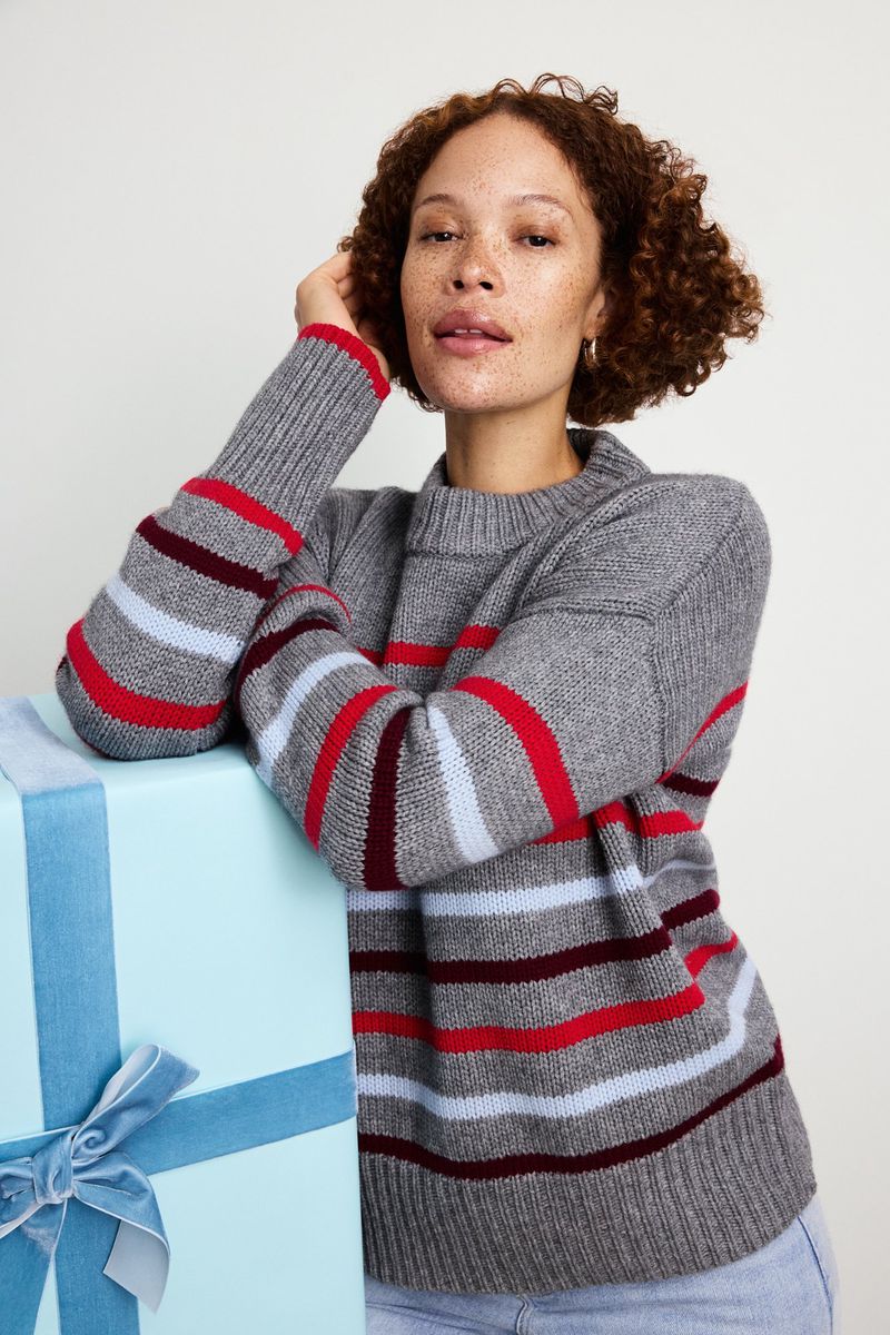 Adult woman with curly hair poses next to a wrapped gift with a blue ribbon. She wears a grey striped sweater in a studio setting.