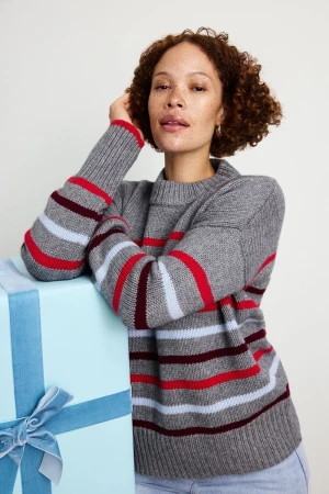 Adult woman with curly hair poses next to a wrapped gift with a blue ribbon. She wears a grey striped sweater in a studio setting.