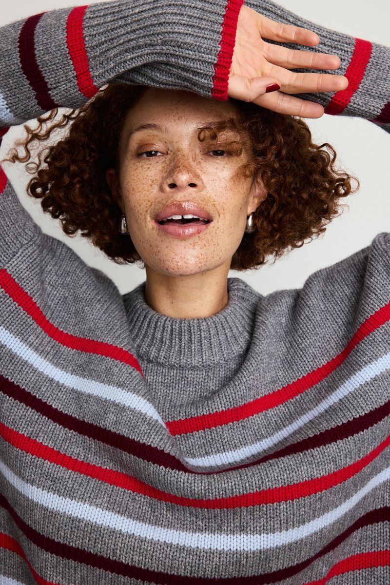 A woman with curly hair poses in a grey striped sweater in a close-up shot, indoors, showcasing beauty and fashion in a studio setting.