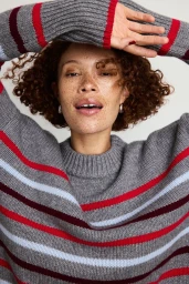 Close-up portrait of a woman with curly brown hair wearing a grey striped sweater against a plain white background.
