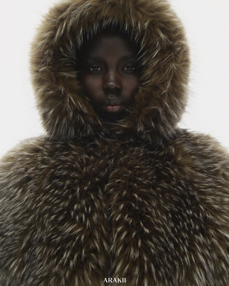 An adult female with dark skin and neutral facial expression posing in a hooded fur coat against a white background in a studio photograph.