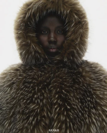An adult female with dark skin and neutral facial expression posing in a hooded fur coat against a white background in a studio photograph.