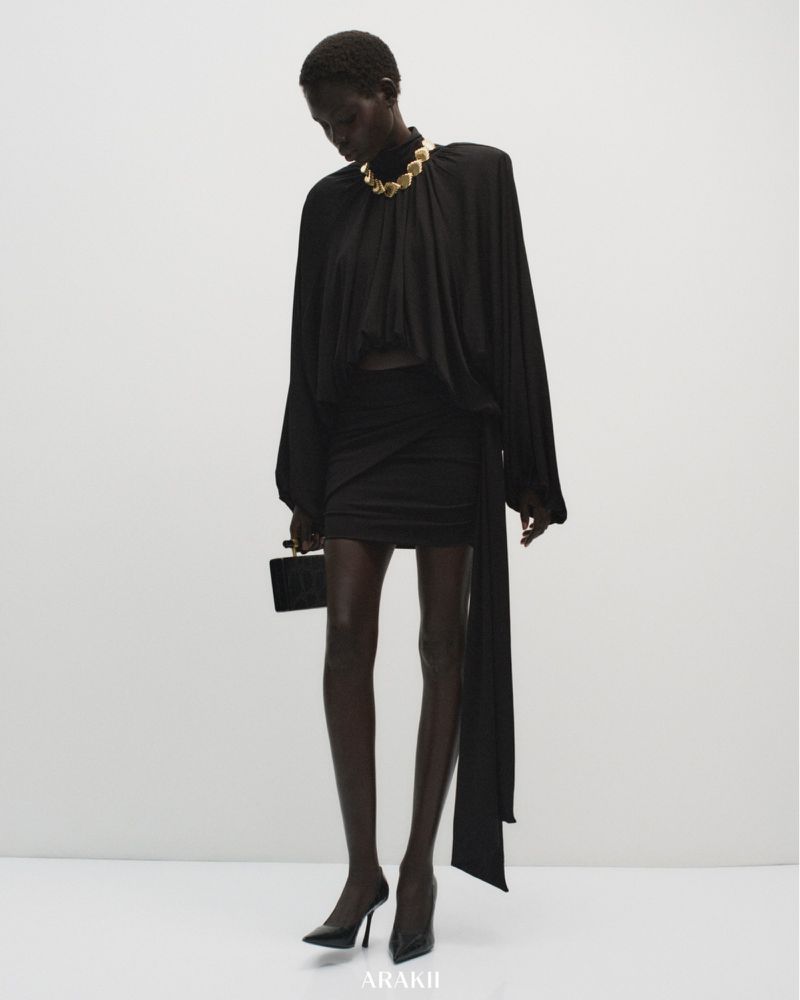 Fashion shot of a Black female model in a draped black top and skirt, accessorized with a gold choker, holding a black clutch against a white backdrop.