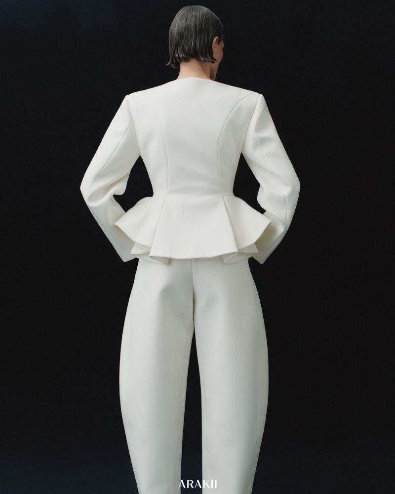 Back view of a fashion model wearing a white peplum blazer and balloon-style pants against a dark backdrop in a studio shot.