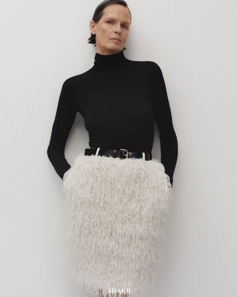 Fashion shot of a mature woman wearing a black turtleneck and a white, textured skirt with a black belt, posing indoors against a white wall.