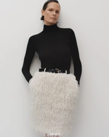 Fashion shot of a mature woman wearing a black turtleneck and a white, textured skirt with a black belt, posing indoors against a white wall.