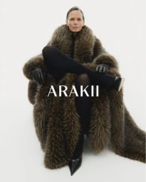 Model seated in a large brown faux fur coat wearing black tights and leather gloves against a white background