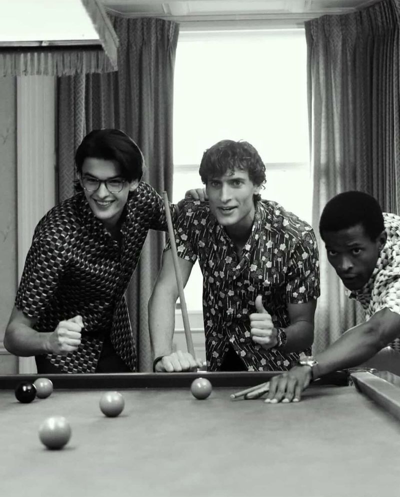 Three young men in patterned shirts gather around a pool table with billiard balls in an indoor setting; monochrome image.