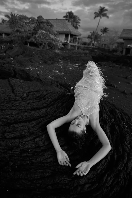 Monochrome fashion shot of a woman in a sequined dress lying on black lava rocks near a tropical resort with palm trees.
