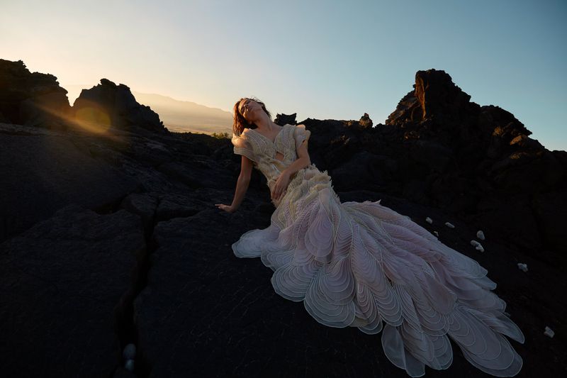 Woman in an elaborate flowing gown poses dramatically on a rocky landscape at sunset, creating a striking fashion editorial image.