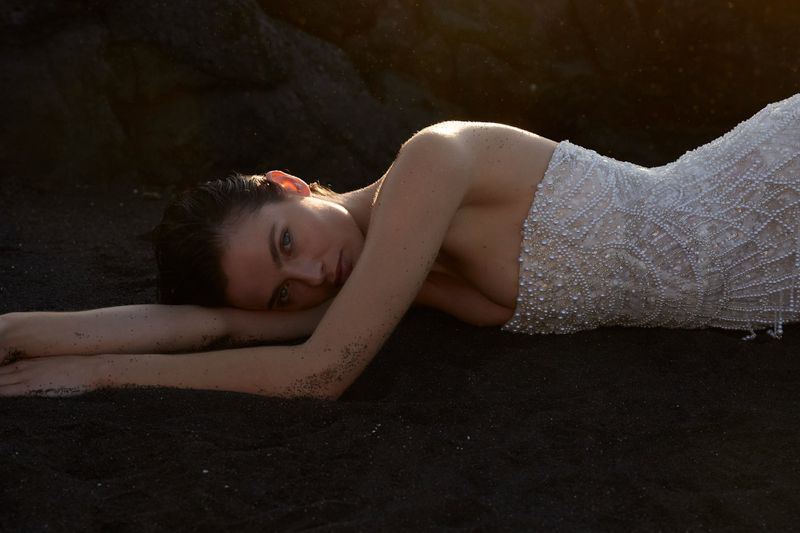 Woman with wet hair lying on black sand wearing a beaded gown, gazing at the camera in an outdoor setting with rocky background.