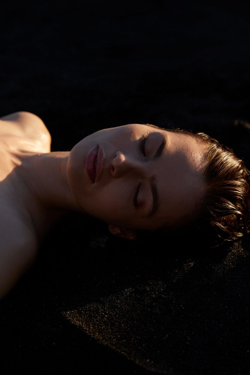 Close-up of a fair-skinned woman lying down with her eyes closed, bathed in warm light against a dark backdrop, portrait orientation.
