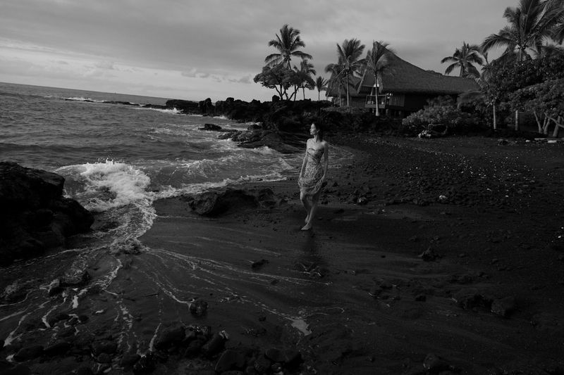 Black and white photo of a woman in a dress walking on a black sand beach with waves crashing, palm trees, and a thatched-roof structure.