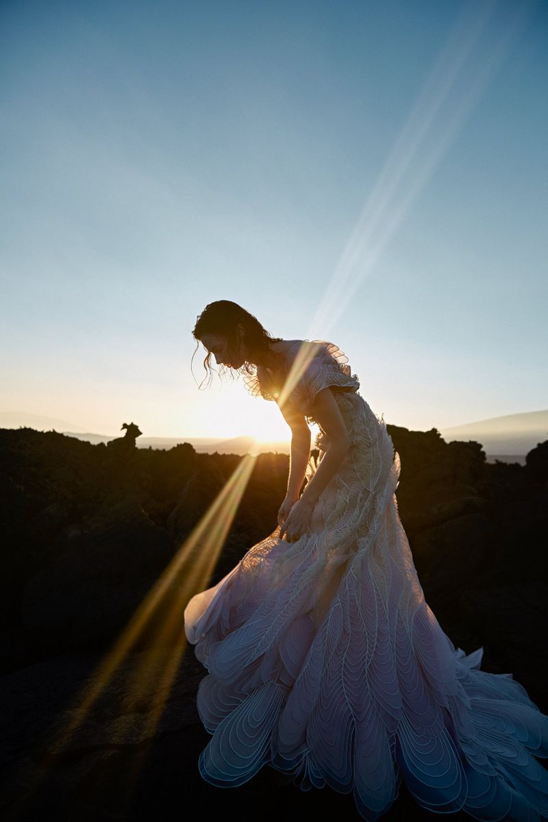 Woman in flowing gown in outdoor landscape at sunset. Low angle captures the subject against the light blue sky and warm sunlight.