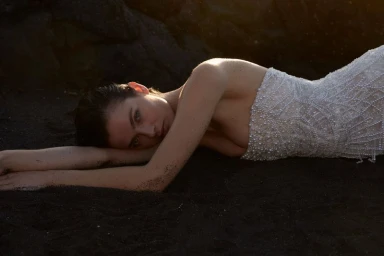 Model Moa Aberg lying on a dark sandy beach wearing a beaded white gown, captured by Sylve Colless for The Lane project.