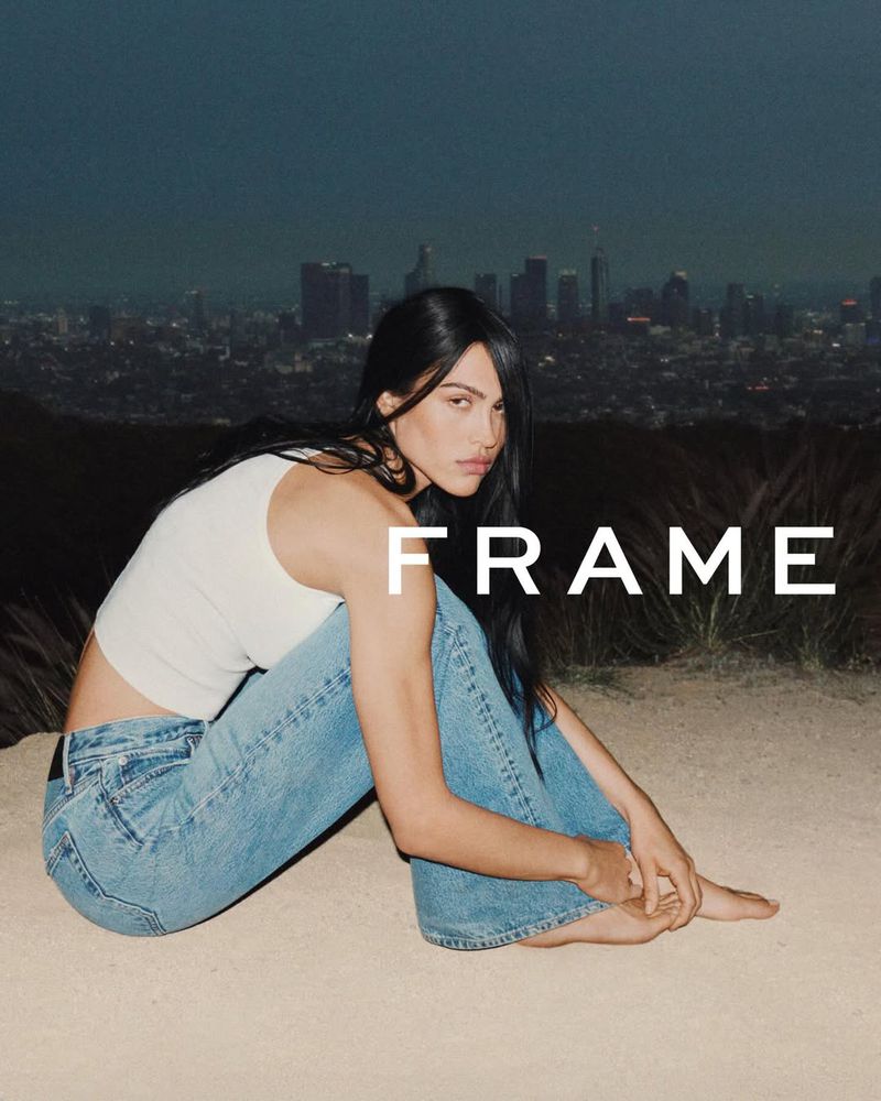 Woman sits barefoot in jeans and a white tank top against an urban backdrop in an outdoor fashion editorial photoshoot.