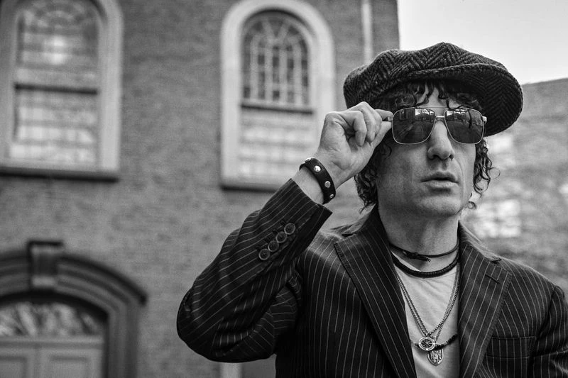 Black and white portrait of a man in a tweed cap and pinstripe suit, adjusting sunglasses against a blurred building backdrop.