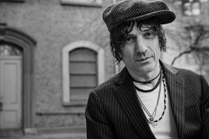 Black and white portrait of an adult male with curly hair, wearing a tweed cap and pinstripe jacket, standing in an urban environment.