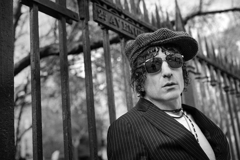 Black and white portrait of adult male in coat, hat, and sunglasses posing in front of metal fence on urban street.