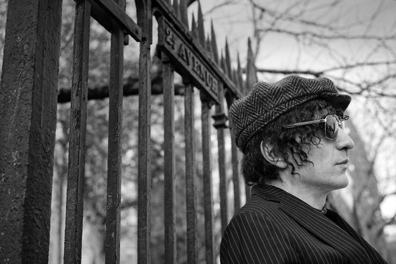 Monochrome portrait of an adult male with sunglasses and cap, standing near a wrought-iron fence with architectural details.