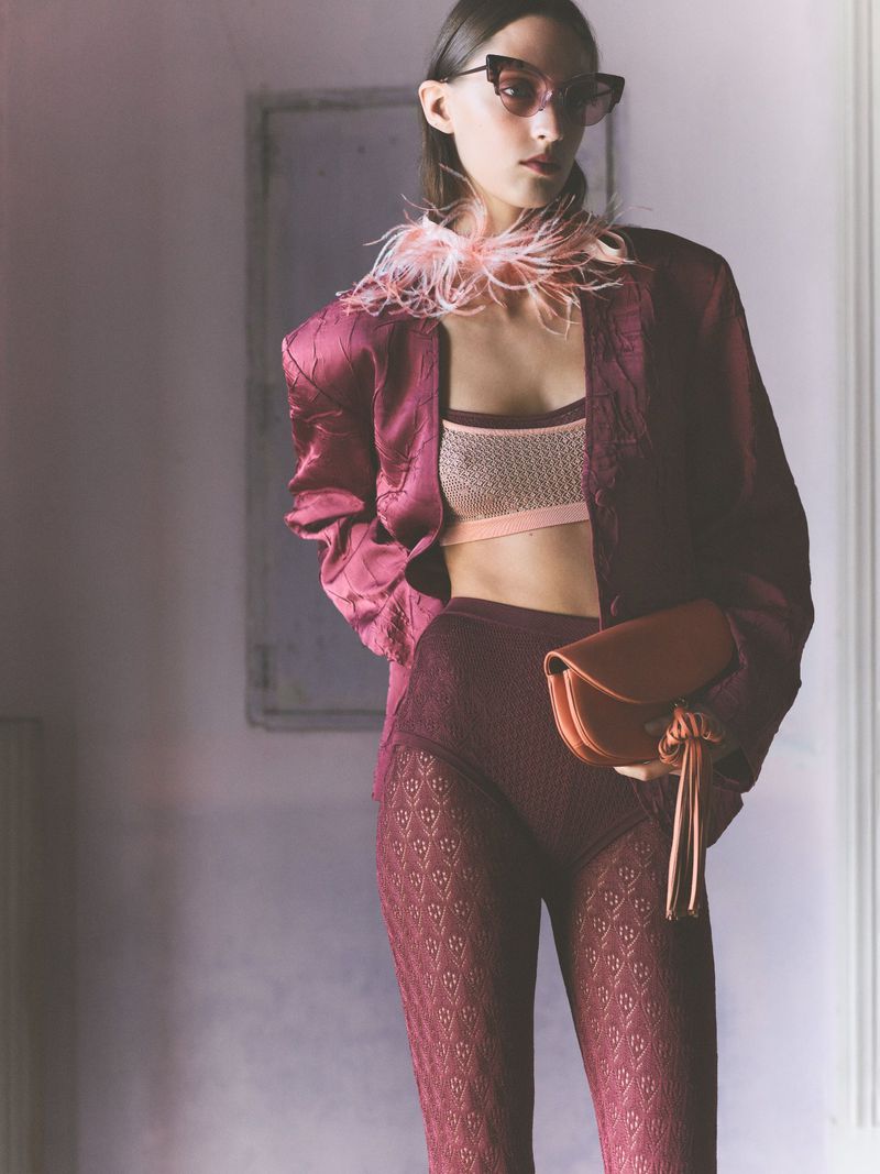 A fashion photo of an adult female model wearing sunglasses and a burgundy outfit, holding a purse, posing in an indoor setting.