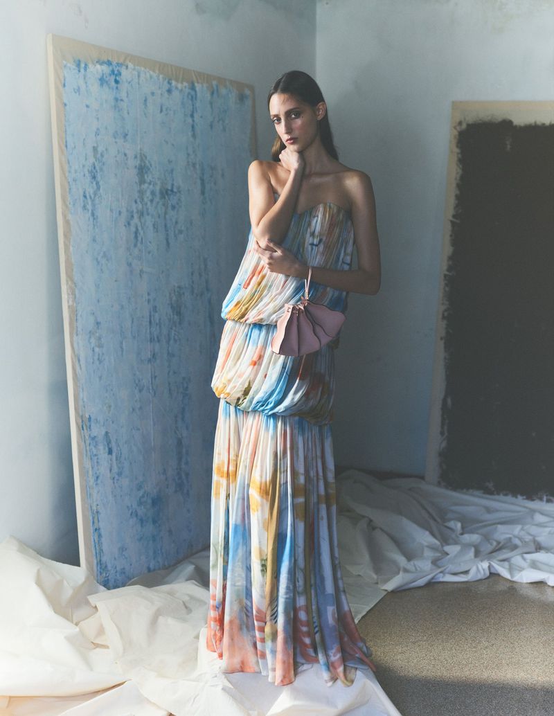 A woman wearing a long strapless dress poses indoors with artwork. The dress has a watercolor-like pattern and a pink purse.