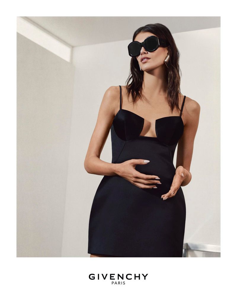 Elegant woman poses indoors wearing a black mini dress, oversized sunglasses, and silver hoop earrings in a fashion editorial.