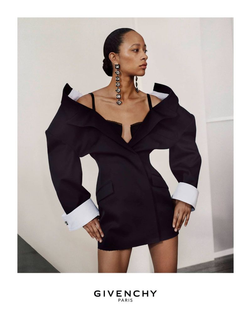Fashion model wearing a black dress and long earrings in a studio, posing against a neutral background in a portrait shot.