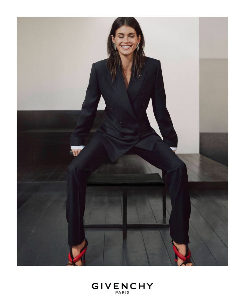 Elegant woman in a black double-breasted suit poses seated in a studio. She wears red heeled sandals with a black wood floor.