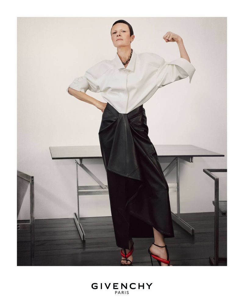 Editorial fashion portrait of a stylish woman in a white button-down shirt and black skirt flexing in a studio setting.