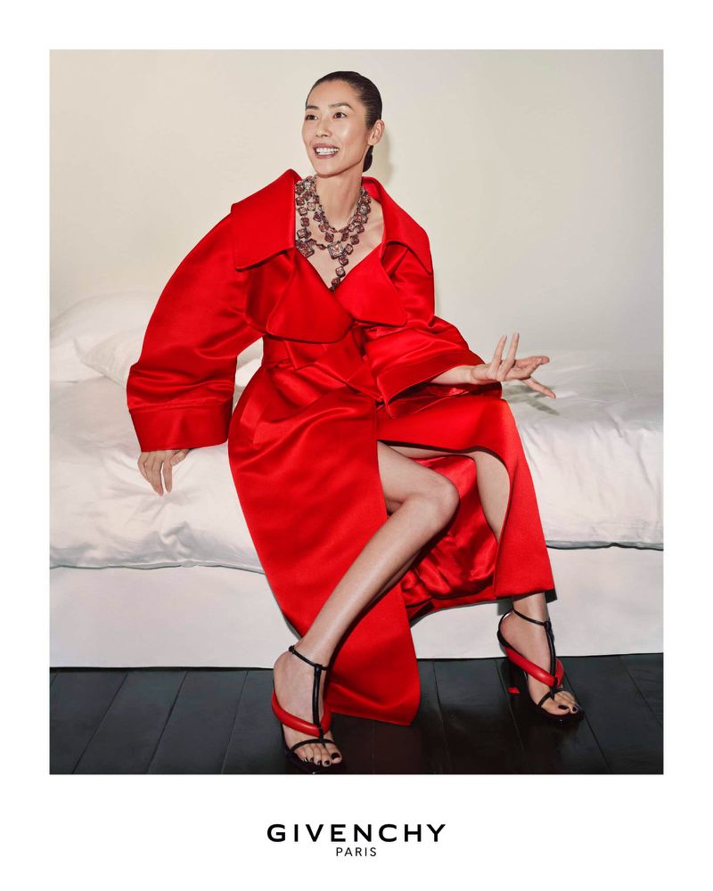 Model Liu Wen wearing a vibrant red coat and statement necklace seated on a white bed for the Givenchy Summer 2026 campaign.