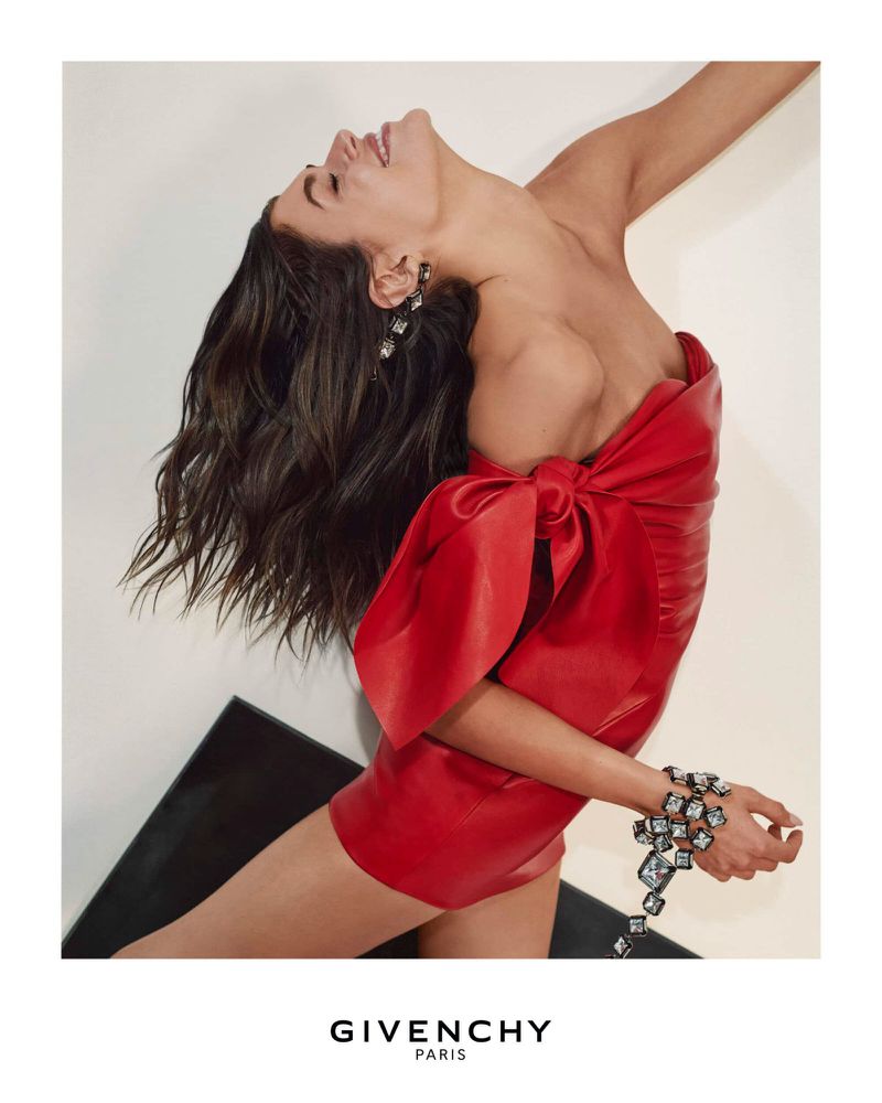 Fashion editorial shot of a woman wearing a red strapless dress and sparkling jewelry, leaning back in a studio with soft lighting.