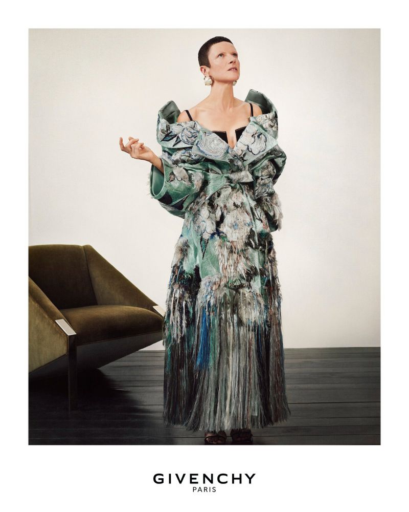 Fashion shot of a woman in an avant-garde gown with floral patterns, posing near a modern armchair against a neutral backdrop.