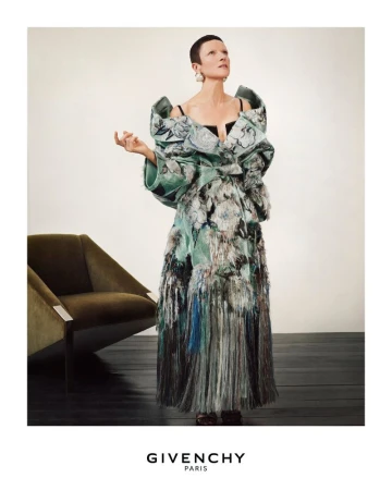 Fashion shot of a woman in an avant-garde gown with floral patterns, posing near a modern armchair against a neutral backdrop.