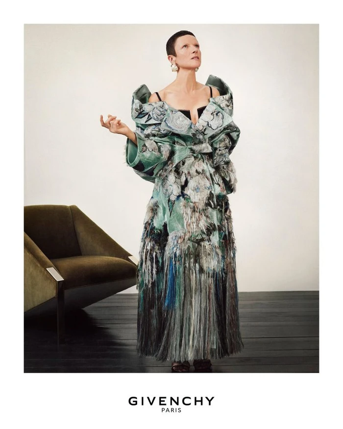 Model with short dark hair in a structured green floral coat standing against a white wall next to a green armchair.