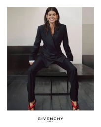 Model wearing a dark tailored pantsuit seated on a stool against a minimalist interior background with Givenchy Paris branding.