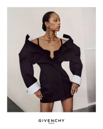 Model in a structured black mini dress with exaggerated shoulders and white cuffs, wearing long crystal earrings against a plain white studio background.