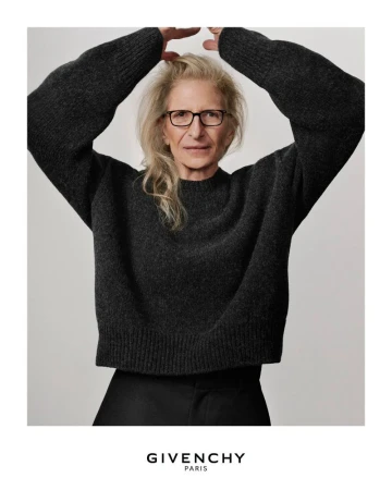Annie Leibovitz wearing a dark grey sweater with arms raised against a plain neutral background for Givenchy