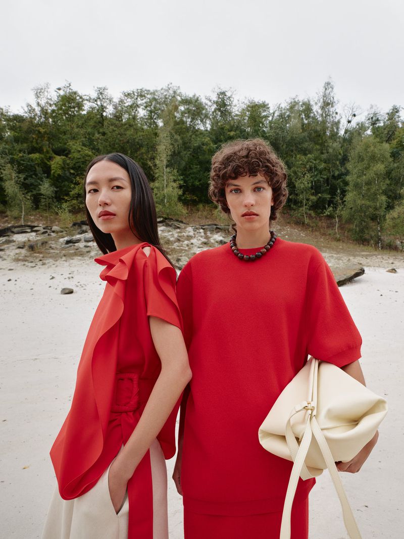 Two fashion models in red tops and dresses pose on a sandy surface against a green forest backdrop in a medium outdoor shot.