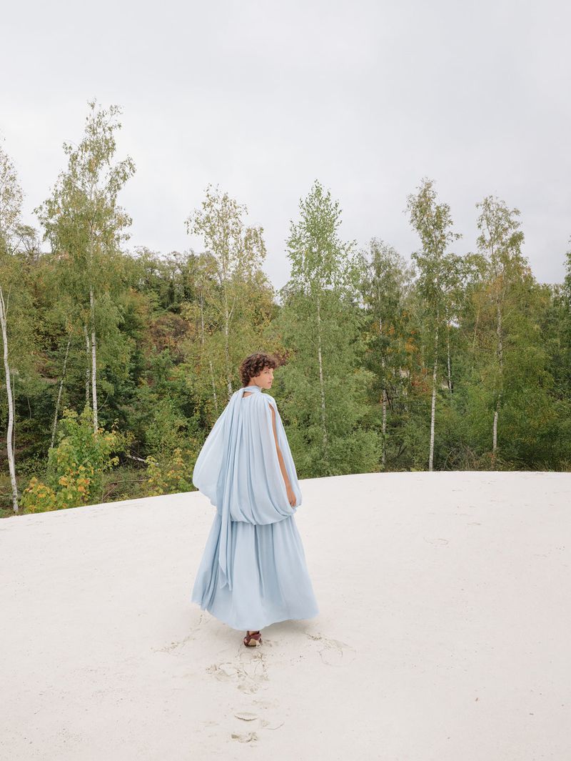 Adult female in long flowing blue dress on light-colored sand, photographed outdoors in a forest clearing on an overcast day.