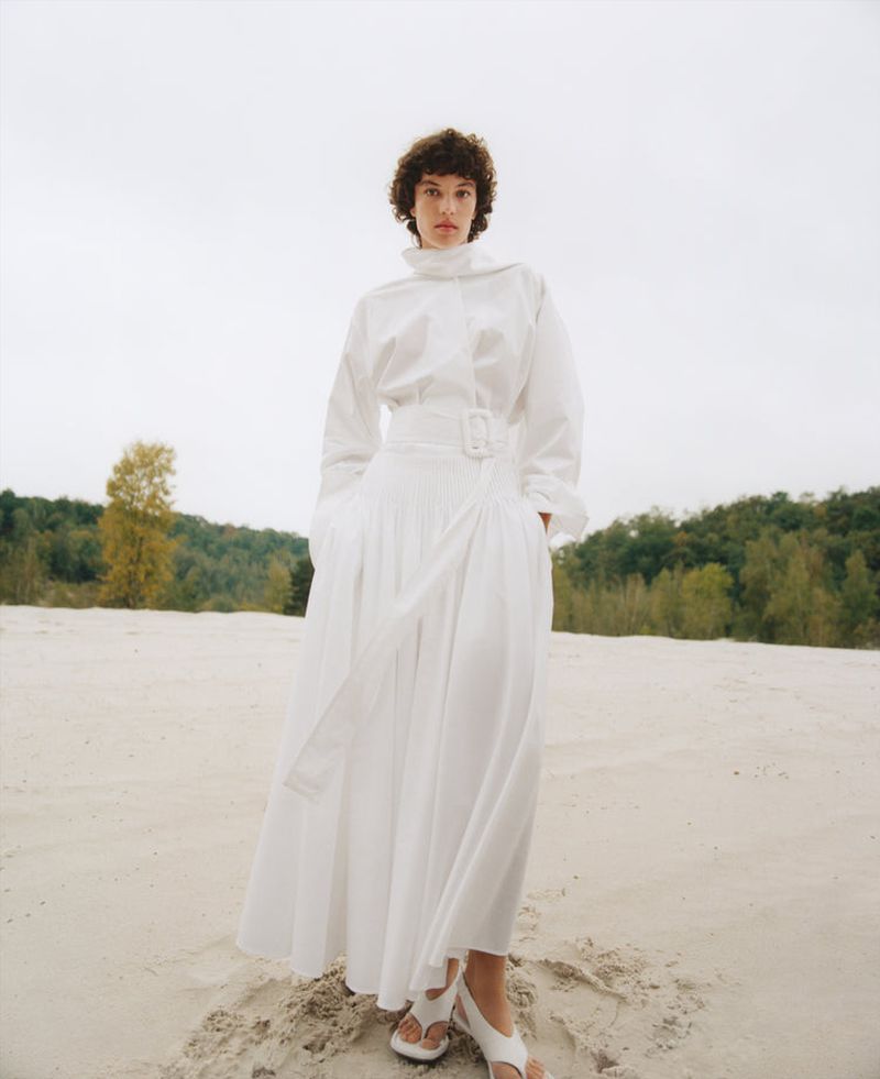 A woman in a white, long-sleeved top and skirt with a white belt stands on a sandy beach, with trees in the background and a cloudy sky.