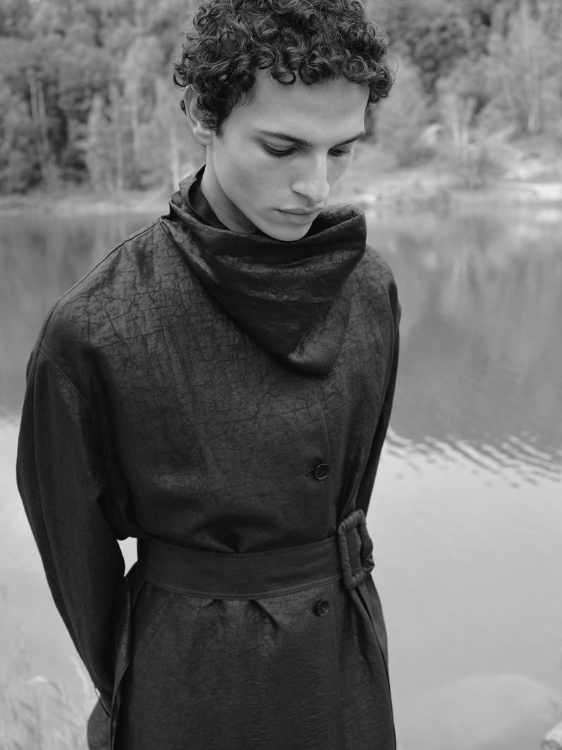 Monochrome portrait of a young adult man with curly hair wearing a dark belted coat, set against a blurred water and forest background.