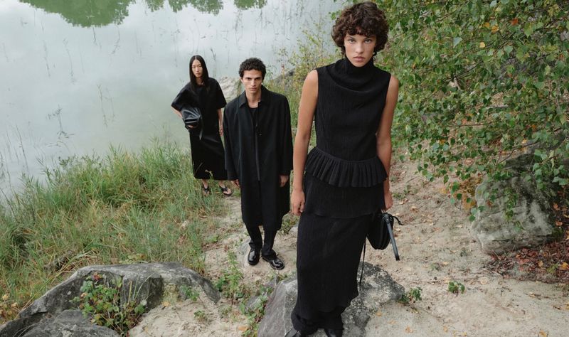 Three fashion models in black outfits pose outdoors near a lake; two women and one man showcase modern, minimalist style in nature.