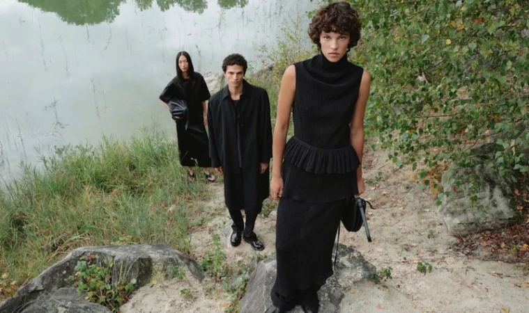 Three fashion models in black outfits pose outdoors near a lake; two women and one man showcase modern, minimalist style in nature.