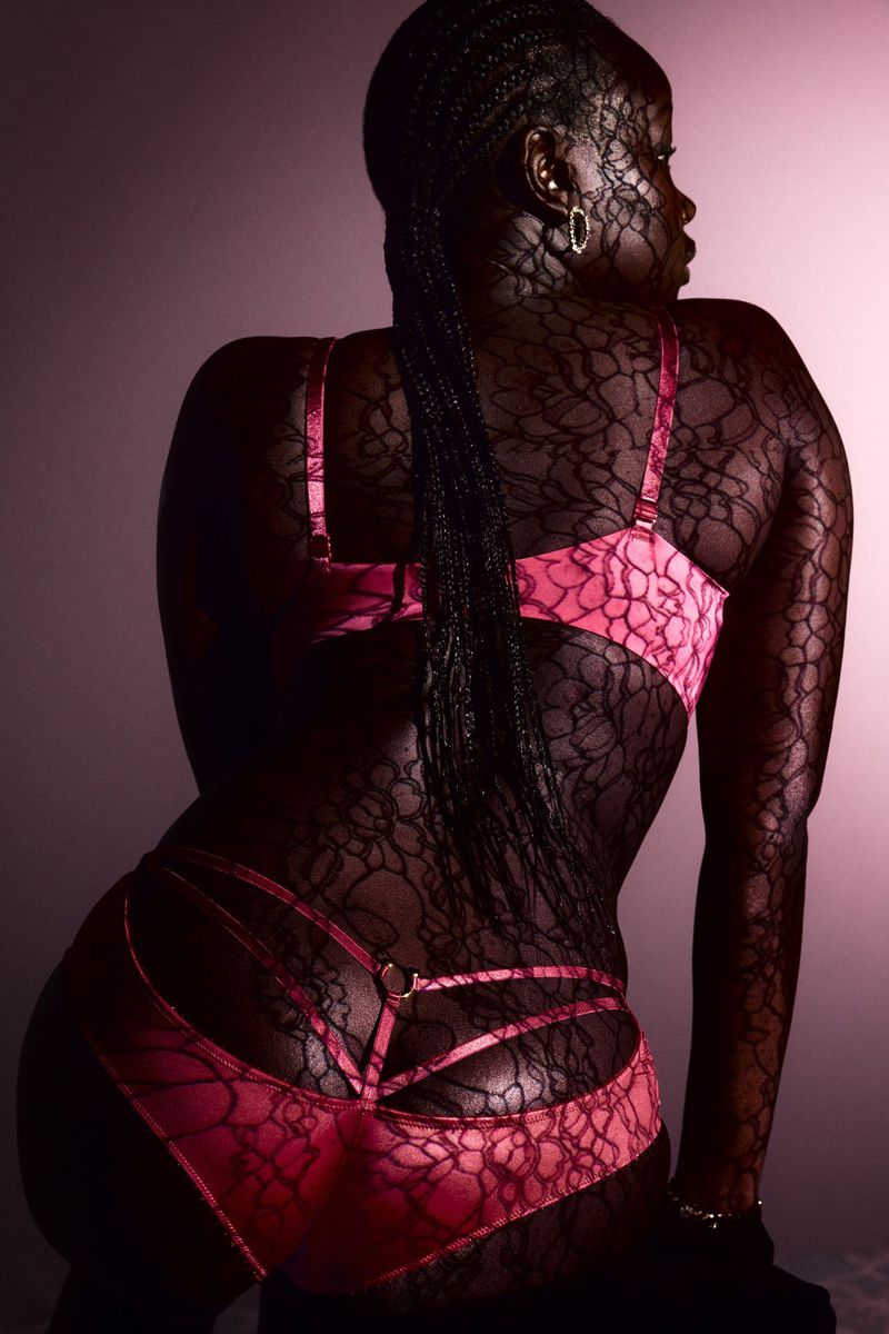 Back view of a woman with braided hair wearing pink lingerie with black lace pattern against a pale pink backdrop in a studio.