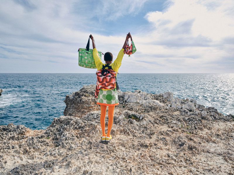 A woman stands on a rocky cliff overlooking the ocean holding shopping bags in the air, styled in bright colors and patterns.