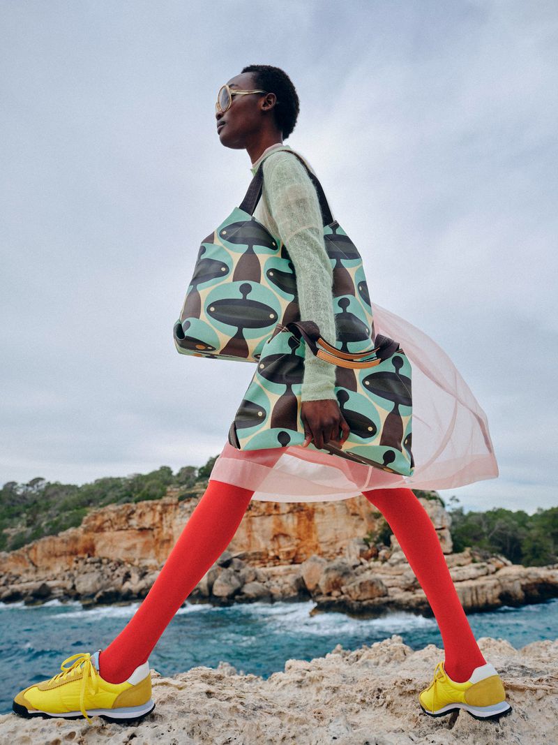 A black female model in a light green sweater, peach skirt, red tights, and yellow sneakers walks with two printed tote bags outdoors.