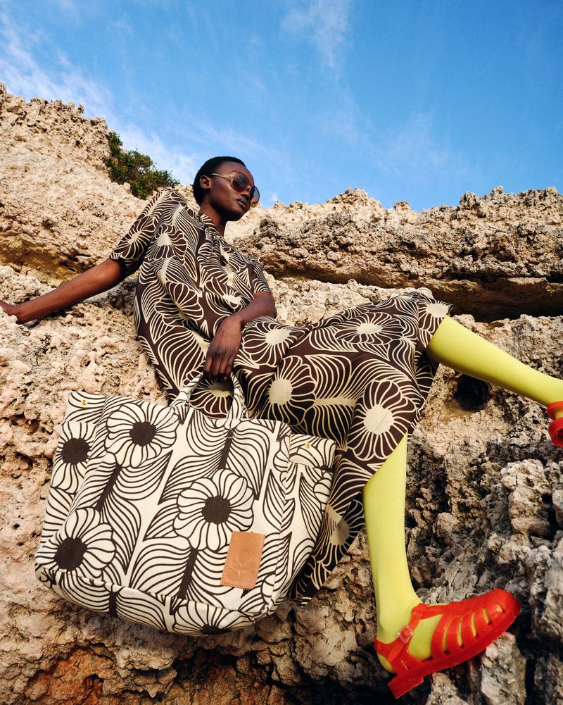 Woman in patterned dress and sunglasses sits on rock formation with matching patterned tote bag against clear blue sky.