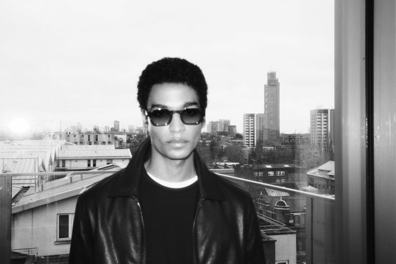 Black and white portrait of a man wearing sunglasses and a leather jacket against a city rooftop background