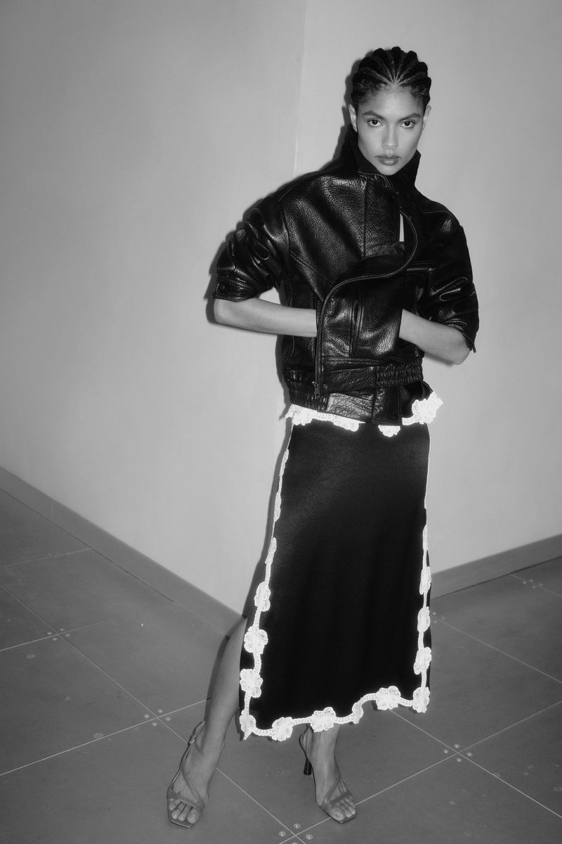 Black and white fashion shot of an adult woman in a black leather jacket and skirt with white trim, posing indoors with arms crossed.